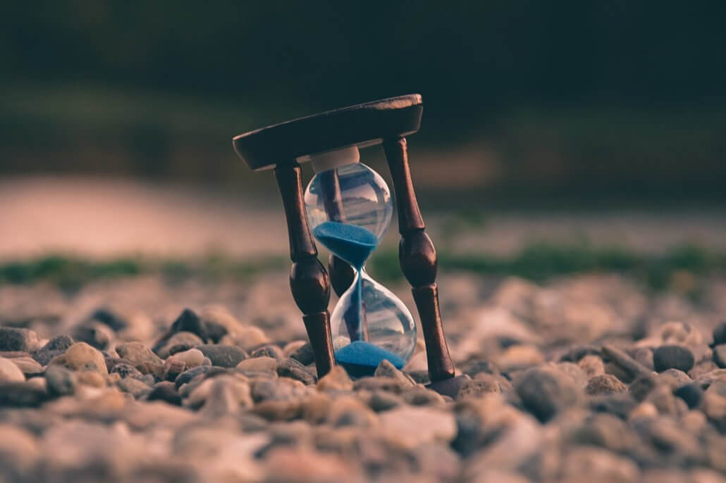 Small hourglass on a pebbled surface with a blurred-out background. In this case, meant to represent time as it relates to estimating web development projects.