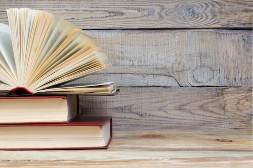 Three books are stacked on a wooden shelf. The top book is open with the pages fanned out.