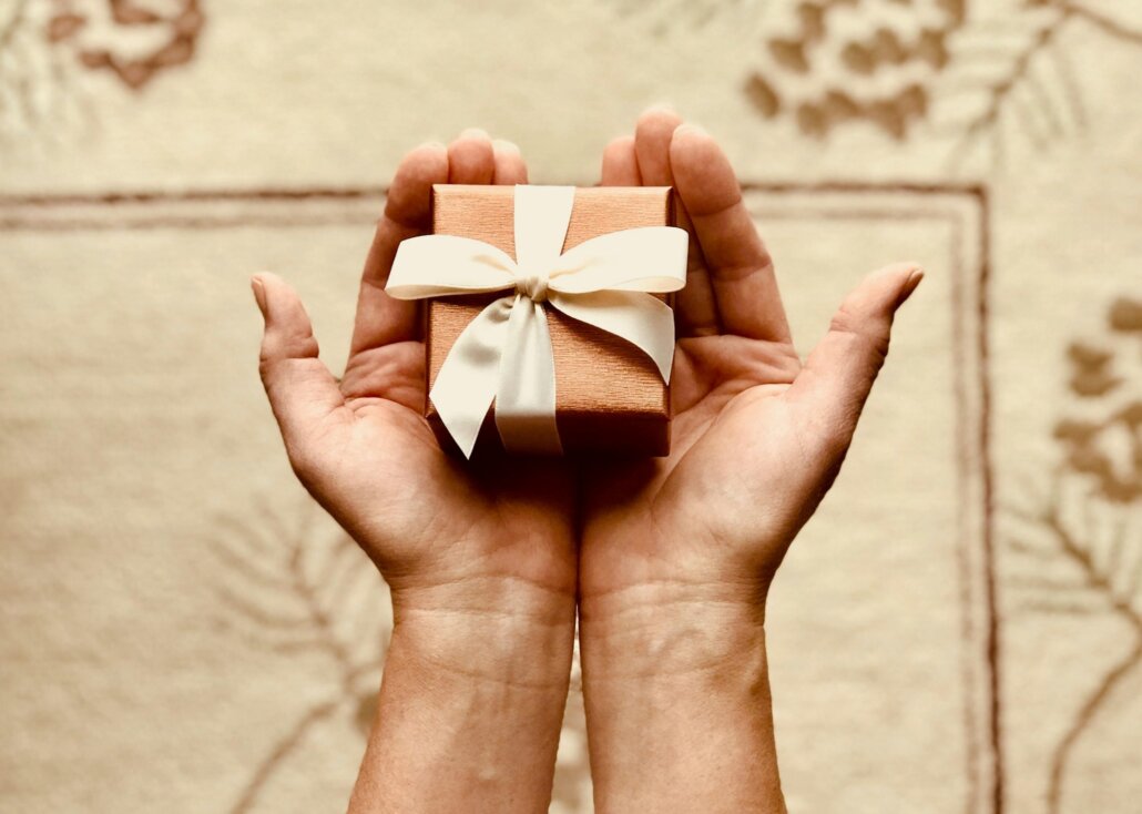 Open hands holding a small box wrapped in brown paper with a white ribbon, representing the act of giving