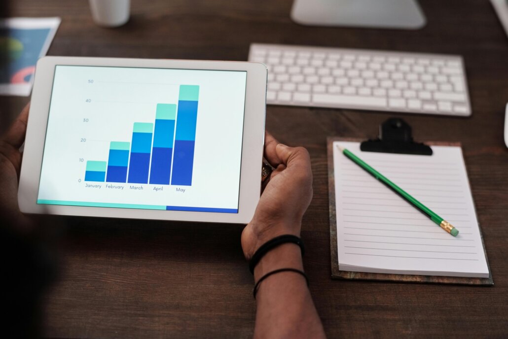 A person holding a white tablet device displaying a bar chart to represent reading an annual report. A computer keyboard, pencil and lined paper can be seen next to the tablet.