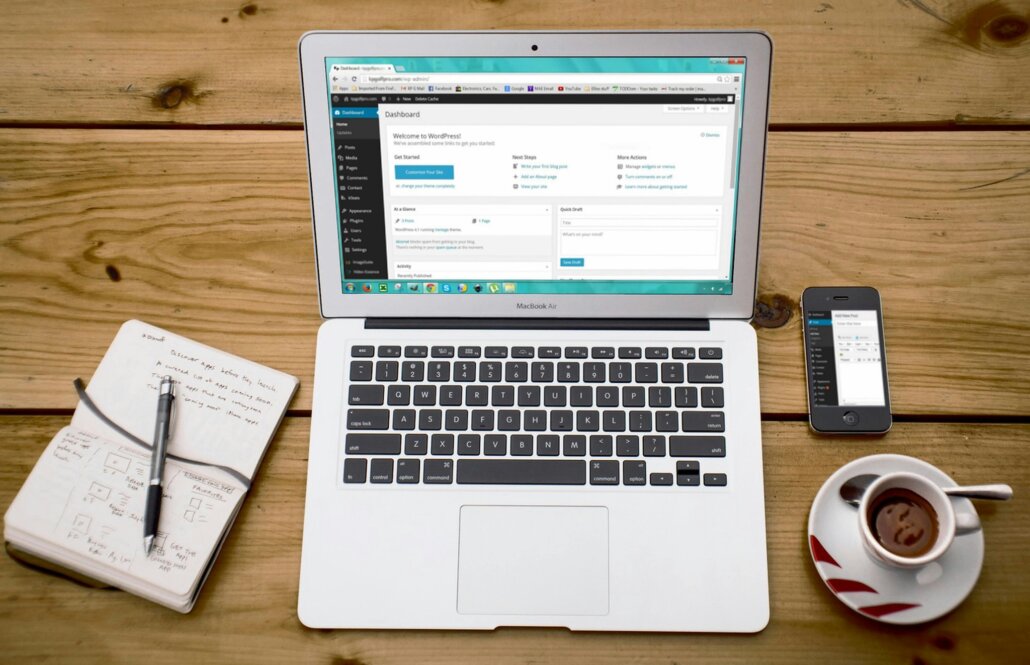 An open laptop computer displays a WordPress dashboard. Next to the computer is an open notebook, a cup of coffee and a mobile phone representing someone's workspace.