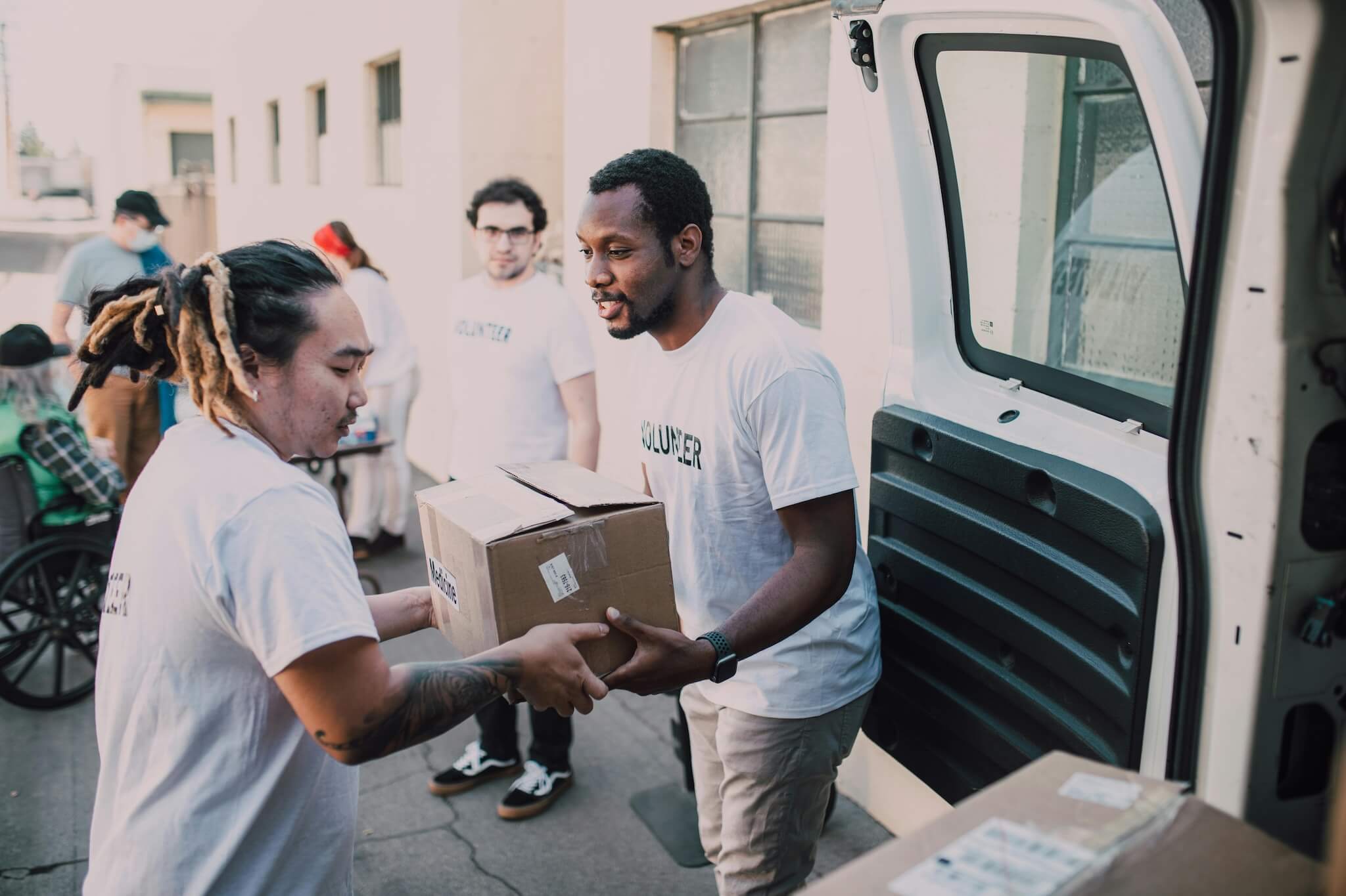Two men standing next to a van with an open door. They are wearing shirts with the word volunteer and passing a cardboard box labelled medicine.
